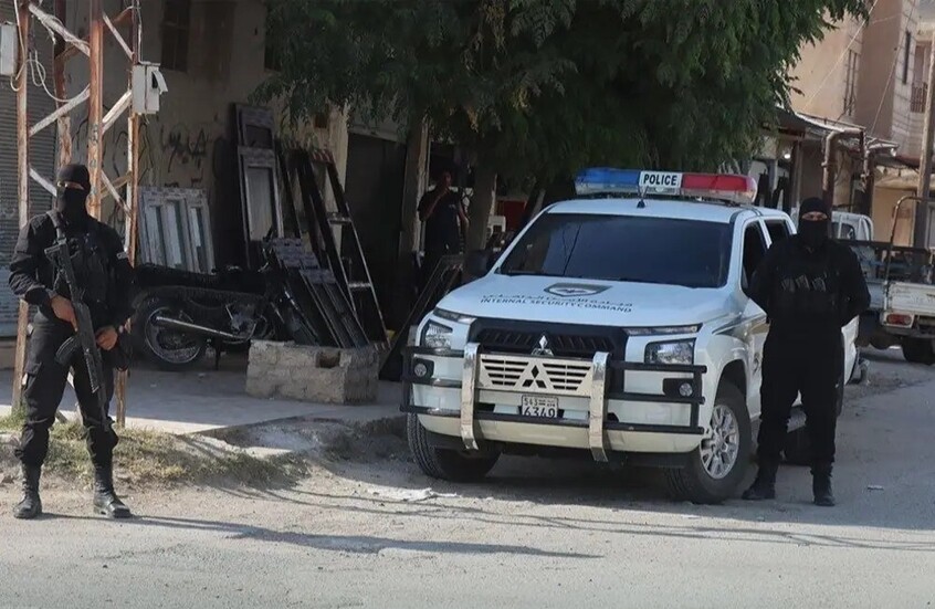 تُظهر الصورة سيارة أمنية سورية بيضاء من طراز "ميتسوبيشي" مخصصة للدوريات ومكتوب عليها باللغتين العربية والإنجليزية "قيادة الأمن الداخلي ويقف بجانبها عناصر أمن ملثمون يرتدون الزي الأسود الكامل ويحمل أحدهم سلاحا رشاشا